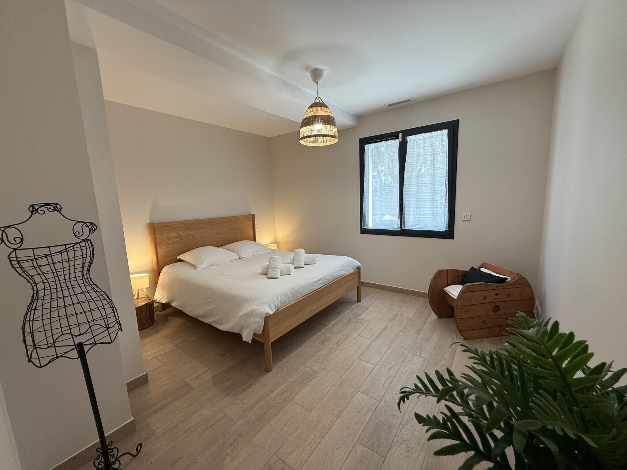 A bedroom with a wooden bed, a hanging light fixture, a window with curtains, a small wooden chest, and a plant.