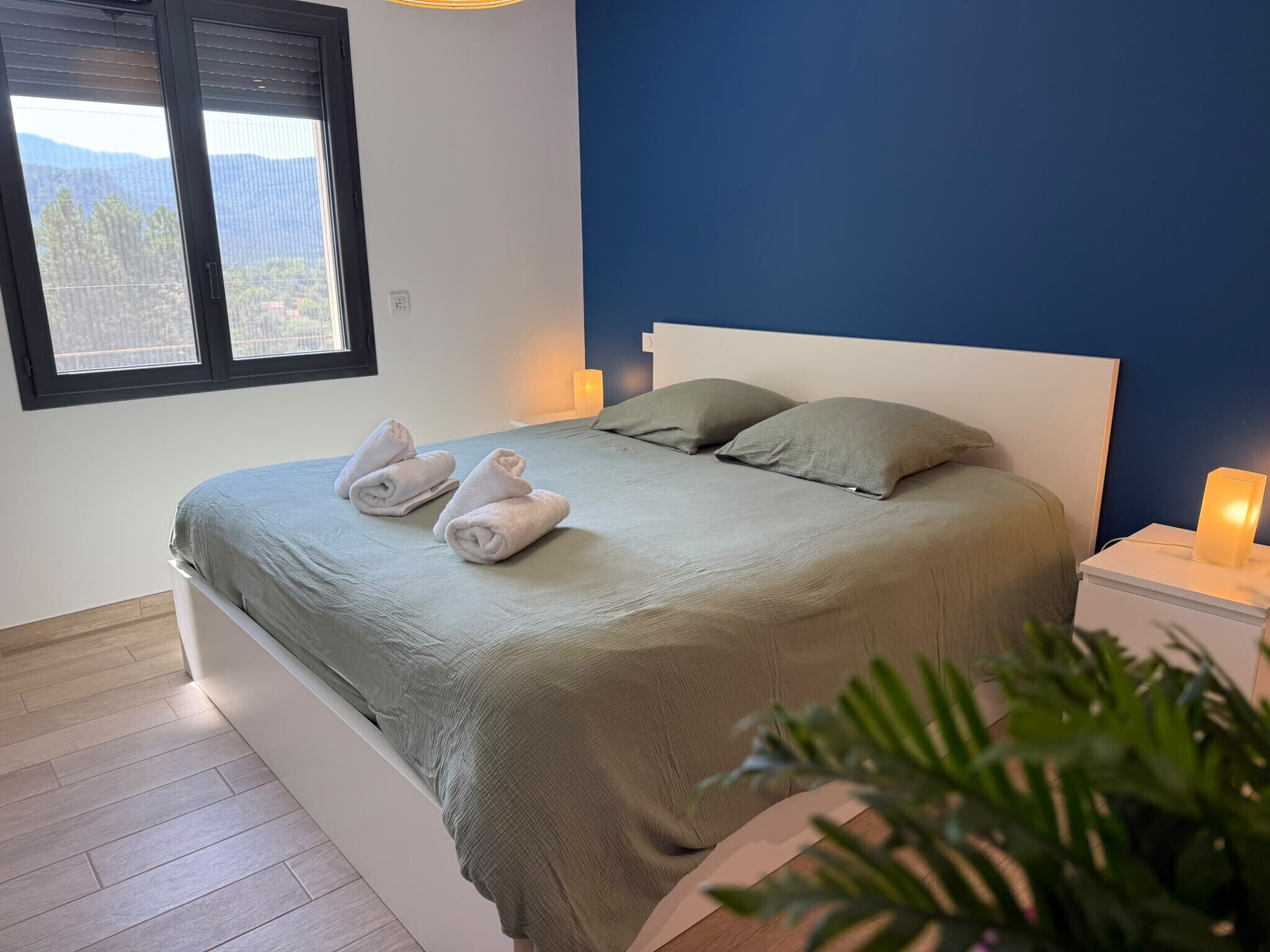 Ein Schlafzimmer mit einem Bett, zwei Kissen, einem Nachttisch mit Lampe und einem Fenster mit Jalousien.