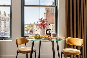 Comedor con una mesa de vidrio, dos sillas, un florero con flores rojas y vistas a la ciudad a través de amplios ventanales.