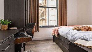 A bedroom with a bed, a desk, a chair, a window with curtains, and a view of buildings outside.