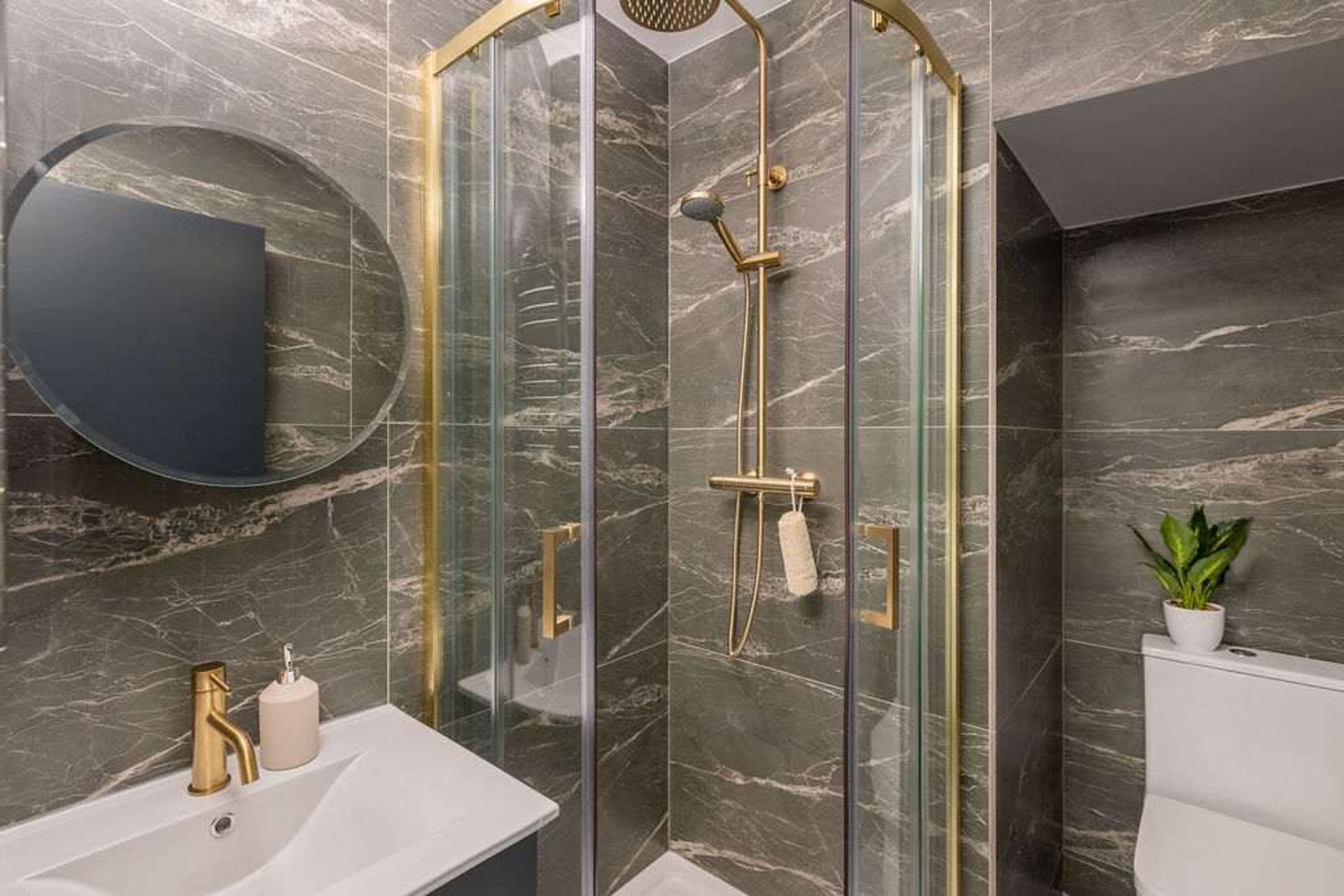 A modern bathroom with a glass-enclosed shower, a round mirror, a white sink, and a potted plant.