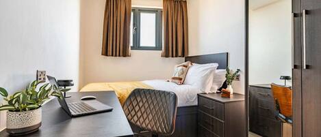A bedroom with a bed, a desk with a laptop, a chair, a dresser, and a window with curtains.
