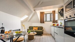 Une cuisine moderne dotée d’un four encastré, un coin repas avec une table en verre et un espace de vie chaleureux avec un canapé et une fenêtre.