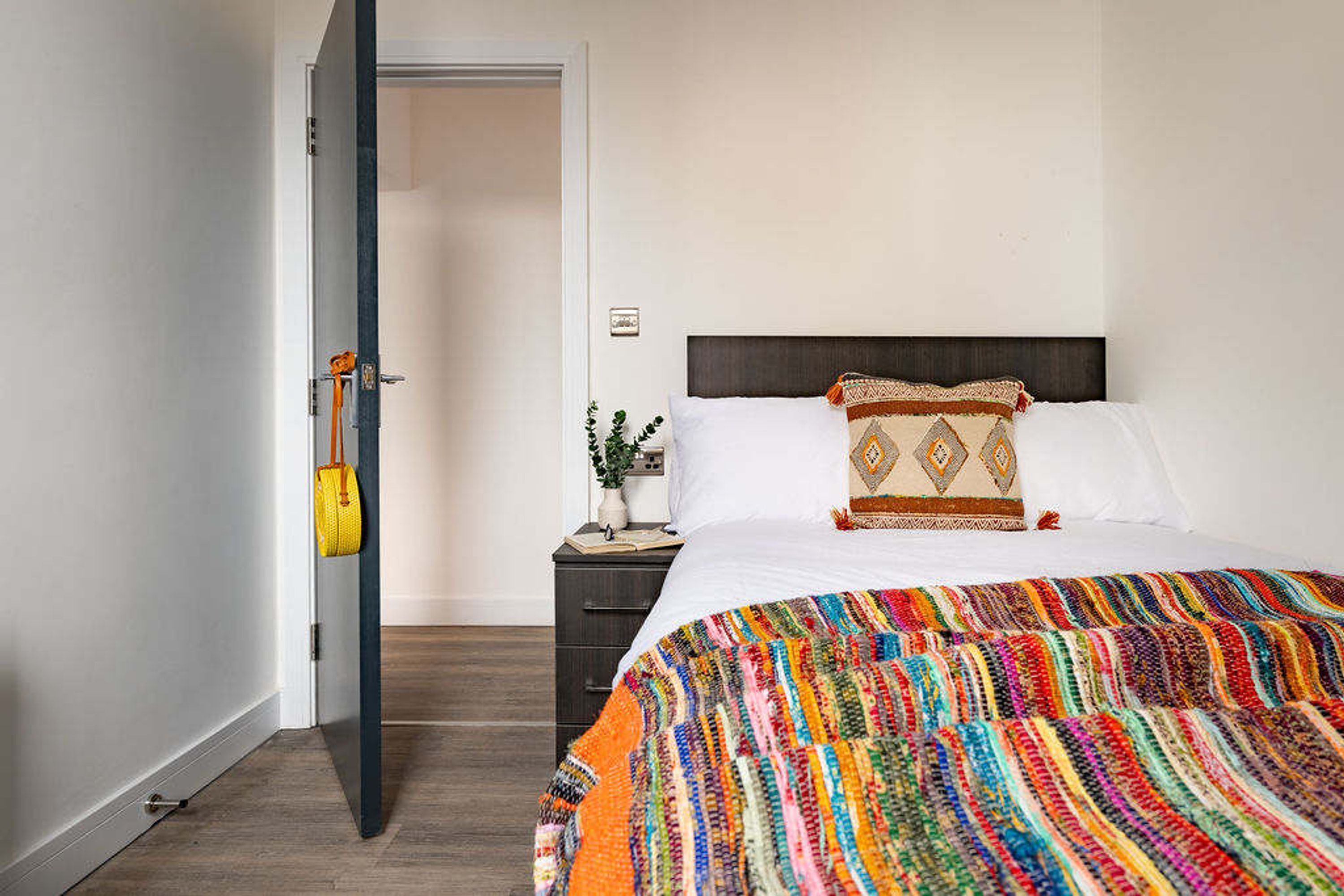 A hotel room with a bed, a nightstand, a door, and a colorful blanket.