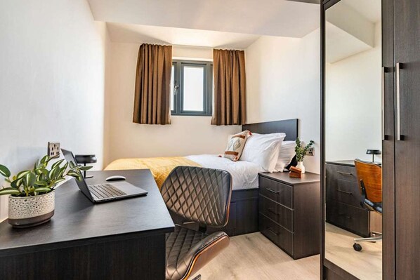 Un dormitorio con cama, escritorio con laptop, silla, cómoda y ventana con cortinas.