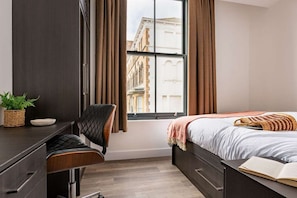 Un dormitorio con cama, escritorio, silla, ventana con cortinas y vista a edificios.