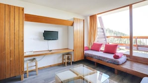 Een moderne kamer met een aan de muur bevestigde televisie, een houten bank met kussens, een glazen salontafel en uitzicht op het groen.