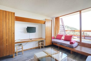 Ein modernes Zimmer mit an der Wand befestigtem Fernseher, einer hölzernen Bank mit Kissen und einem Glastisch, mit Blick ins Freie.
