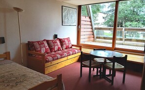 Ein gemütliches Zimmer mit einer Holz-Couch, einem kleinen Tisch und Stühlen. Ein Fenster bietet Blick auf Bäume und eine Holzterrasse.