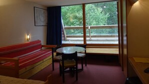 A room with a red sofa, a small table with chairs, a window with a view of greenery, and a wooden railing. - 50m from ski slopes, 1st floor, balcony, tv, 30m², Les Arcs (Les Arcs)