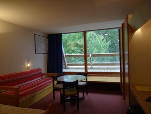 Ein Zimmer mit einem roten Sofa, einem kleinen Tisch mit Stühlen, einem Fenster mit Blick auf Grünflächen und einem Holzgeländer.