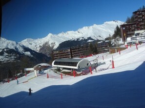 Ein Skigebiet mit schneebedecktem Berg im Hintergrund, einer überdachten Sesselbahnstation und einem mehrstöckigen Gebäude.