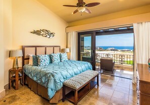 Quarto com uma cama grande, um banco, um ventilador de teto, uma mesa de cabeceira e vista para a praia através de uma porta de vidro.