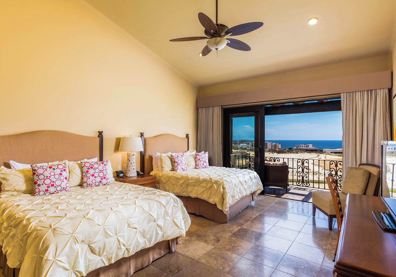 A hotel room with two beds, a ceiling fan, a balcony with a view of the beach, and a desk with a chair.