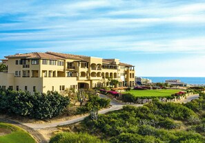 Um hotel grande, de vários andares, com arquitetura mediterrânea, com varandas, um pátio com vegetação e vista para o mar.