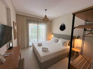 A hotel room with a bed, a desk with a chair, a TV, a hanging light fixture, and a window with curtains. - Darion Residence A5 - Apartament Steps from the Sea (Nikiti)