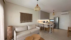 Una sala de estar moderna con sofá, mesa de comedor y sillas, un área de cocina con refrigerador y una lámpara colgante.