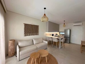 Ein modernes Wohnzimmer mit einer Couch, einem Esstisch und Stühlen, einer Küchenzeile mit Kühlschrank und einer Hängeleuchte.