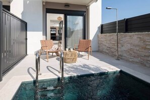 A swimming pool with two wicker chairs and a small round table, a modern building with a glass door, and a stone wall. - Villas Thallo № 4 (Neos Marmaras)
