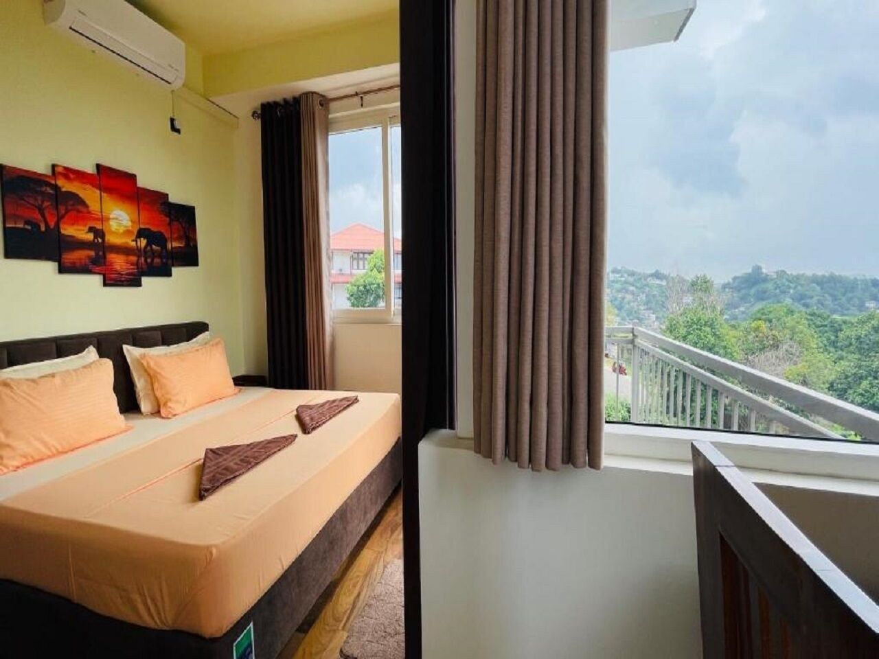 A hotel room with a bed, a painting, a window with curtains, and a balcony with a railing.