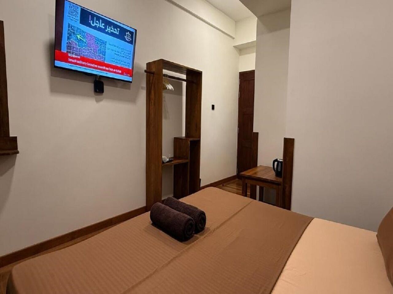 A hotel room with a bed, a television mounted on the wall, a wooden wardrobe, and a small table with a cup.