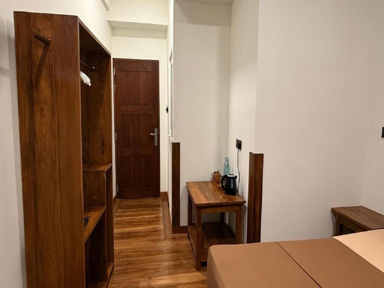 A room with a wooden wardrobe, a desk with a kettle, and a bed.