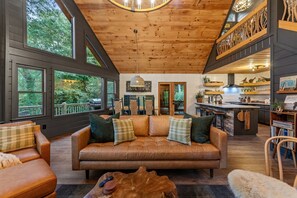 Ein gemütliches Blockhaus-Interieur mit Ledersofa, Holzbalkendecke und großen Fenstern mit Blick auf einen Wald.