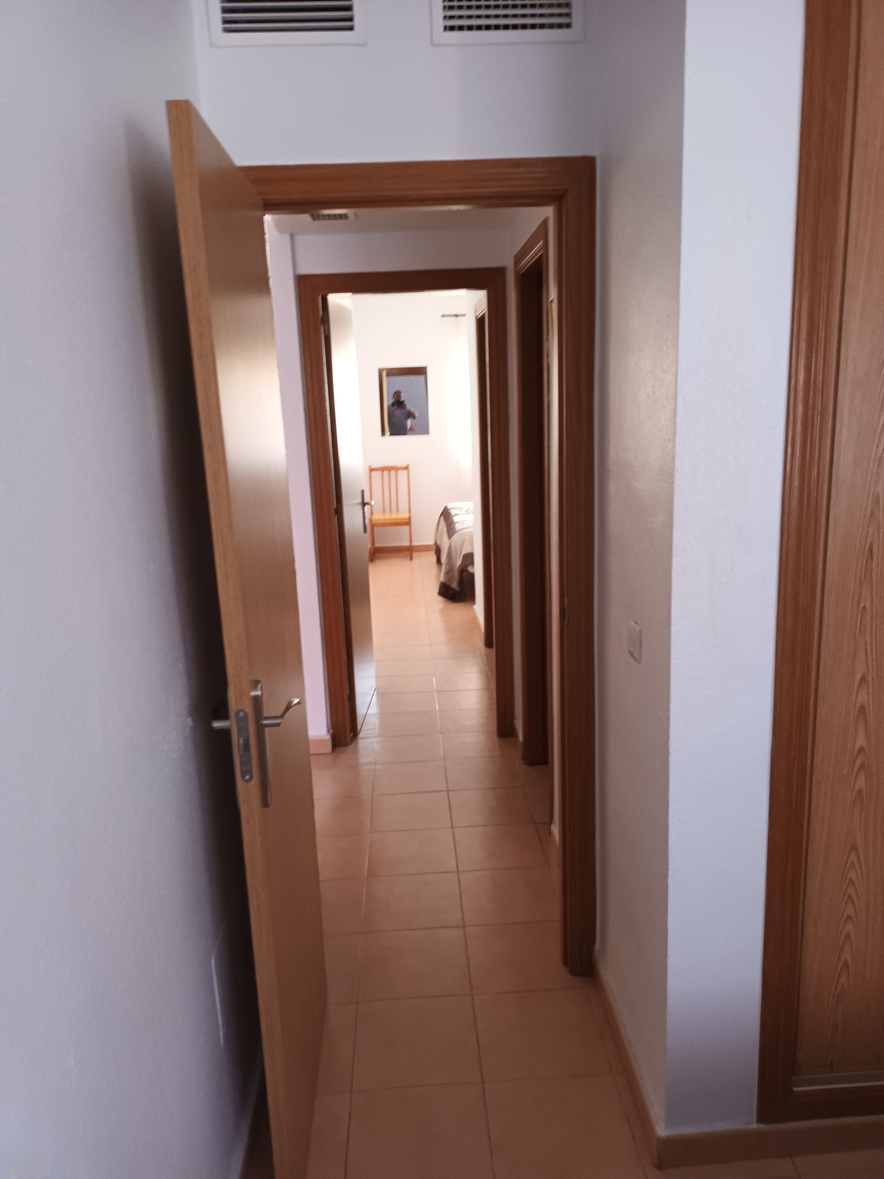 Un couloir avec une porte en bois, une chaise et, au loin, un lit.