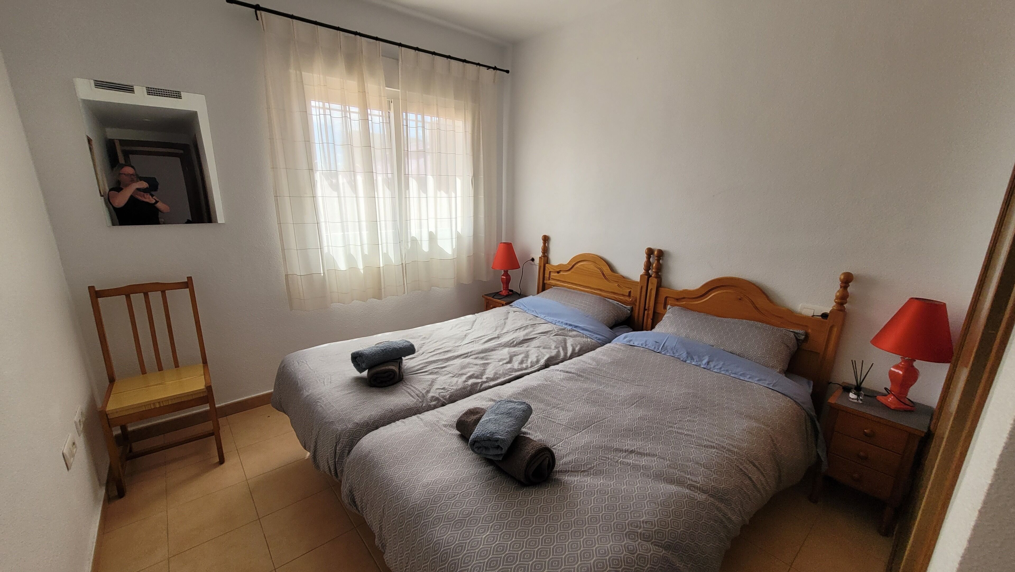 Een slaapkamer met twee bedden, een stoel, een nachtkastje met lampen en een raam met gordijnen.