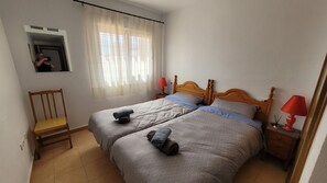 Ein Schlafzimmer mit zwei Betten, einem Sessel, einem Nachttisch mit Lampen und einem Fenster mit Vorhängen.