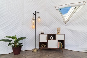 Ein Zimmer mit einer gepolsterten weißen Wand, einer Zimmerpflanze, einer Stehlampe, einer Holzkassette mit Büchern und einem Fenster mit Blick.