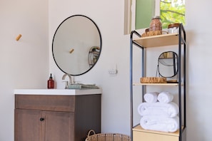 Un baño con un mueble de madera, un espejo redondo, una repisa con toallas y una cesta de mimbre.