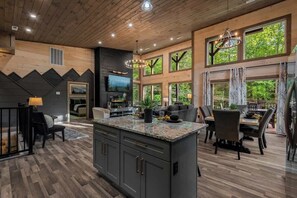 A spacious living area with a large island, dark wood flooring, and a view of greenery through large windows. - 3-Bedroom Gatlinburg Cabin with Indoor Pool & Private Pickleball Court (Gatlinburg)
