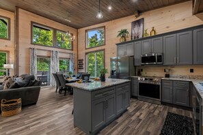 A spacious kitchen with dark cabinets, a large island, and a dining area with a view of trees. - New Lodge in Gatlinburg + Indoor Pool, Hot Tub, Pickleball Court + Sleeps 10 (Gatlinburg)