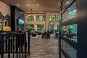 A modern interior with a wooden floor, a TV mounted on the wall, and a large window with a view of trees. - New Lodge in Gatlinburg + Indoor Pool, Hot Tub, Pickleball Court + Sleeps 10 (Gatlinburg)