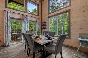 Un coin repas avec une table en bois, des chaises et une vue sur la forêt grâce à de grandes fenêtres.