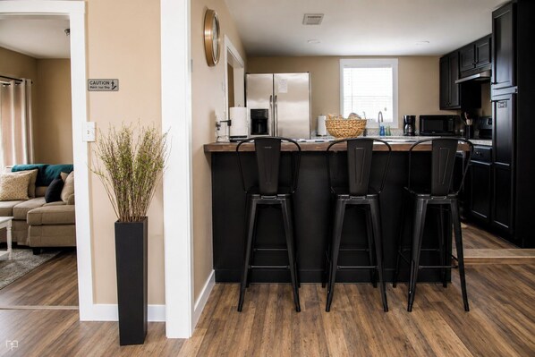 A modern kitchen with a central island, black bar stools, and a dining area with a sofa and decorative plant.