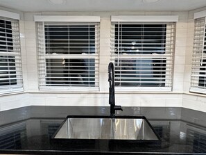 Eine Küche mit schwarzem Arbeitsplatz, weißen Fliesenwänden und einem Fenster mit Jalousien.