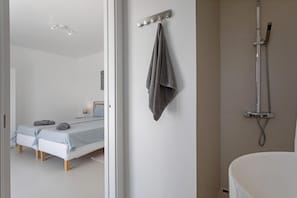 Et moderne badeværelse med bruser, håndklædetørrer og en seng i det tilstødende værelse.