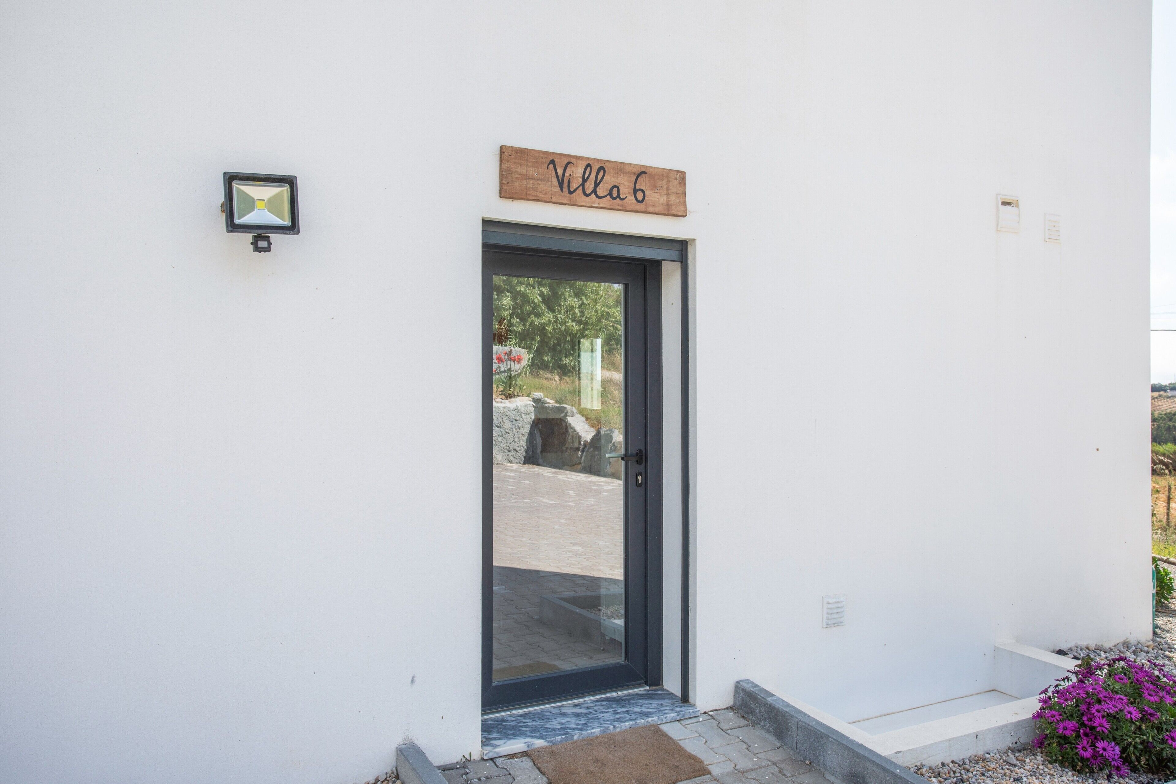 Edificio blanco con puerta etiquetada 'Villa 6', un pequeño espacio exterior con escalones y un parterre de flores.