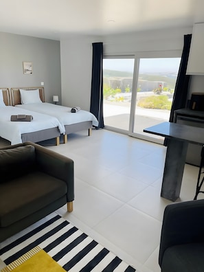 Quarto de hotel moderno com uma cama grande, um sofá, uma mesa de jantar e vista para o exterior.