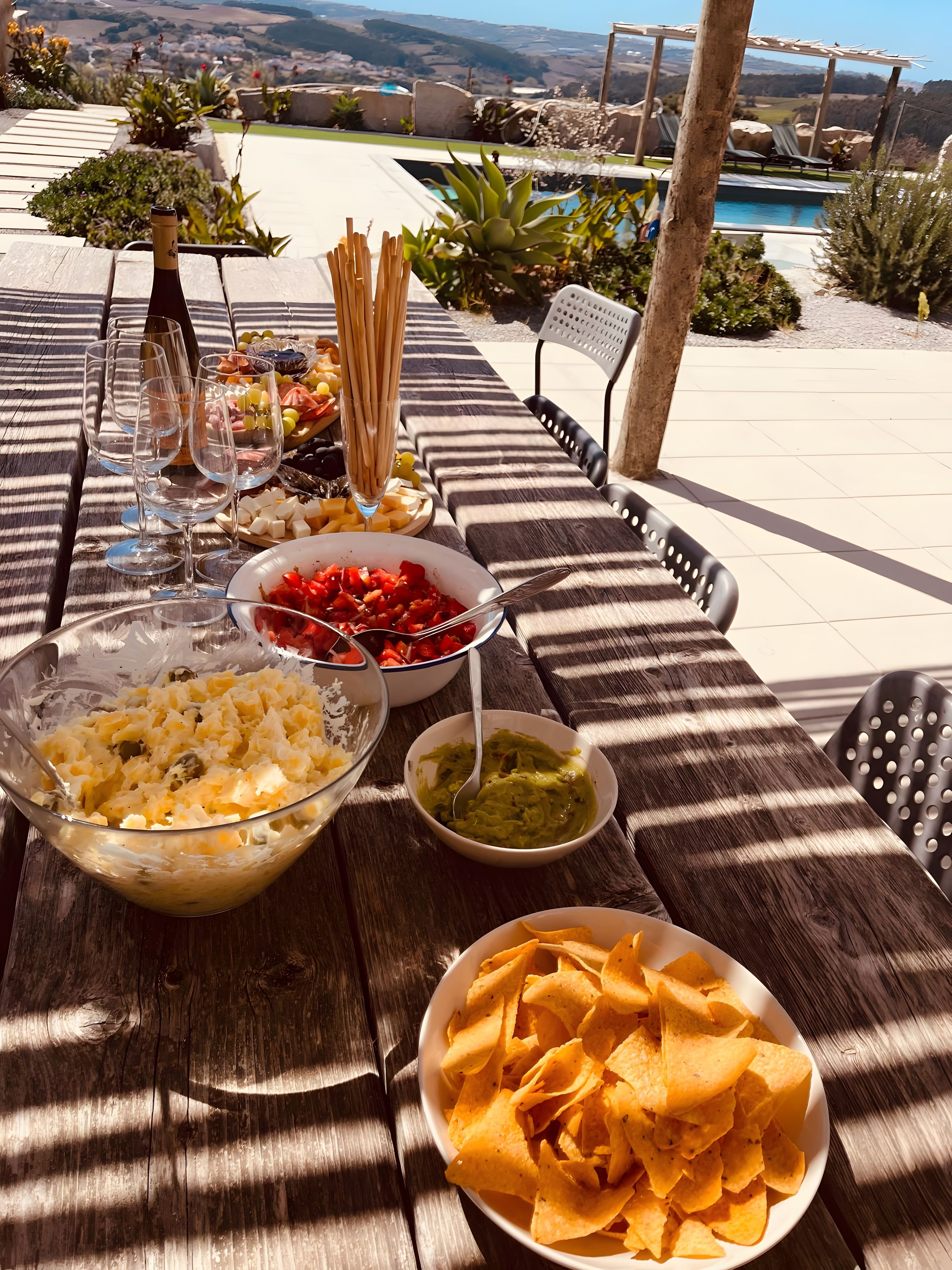 Mesa posta ao ar livre com comida e bebidas, com vista para uma piscina e paisagem.