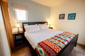 A neatly made bed with white linens and a colorful blanket, a bedside table with a lamp, a window with blinds, and two framed artworks on the wall. - 5 King Rooms, Game Room, Hot Tub, Enclosed Yard, Views (El Prado)