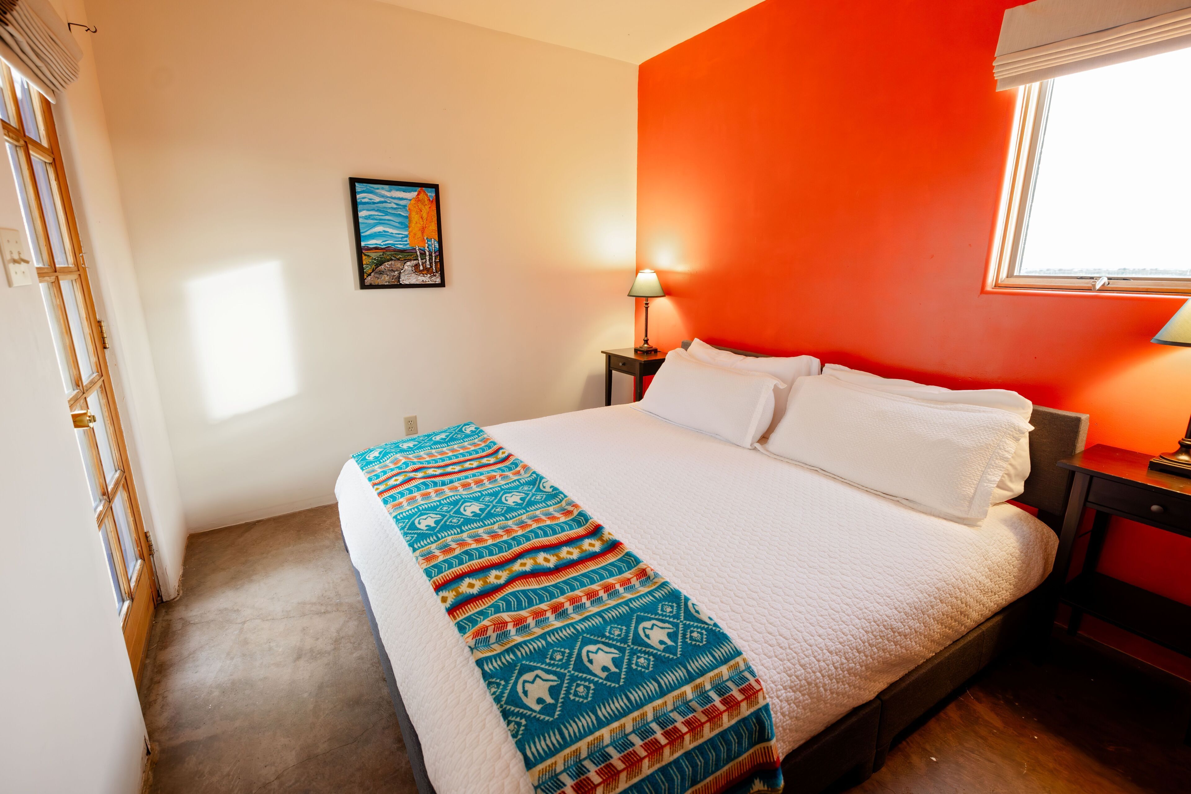 Een slaapkamer met een bed, nachtkastje, lamp, raam en schilderij aan de muur.