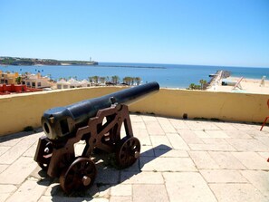 Um canhão numa área pavimentada, com vista para o mar e edifícios ao fundo.