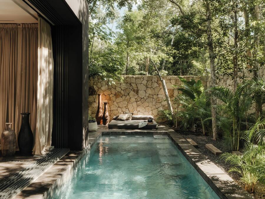 Una piscina con un muro de piedra y follaje, un sofá y dos jarrones.