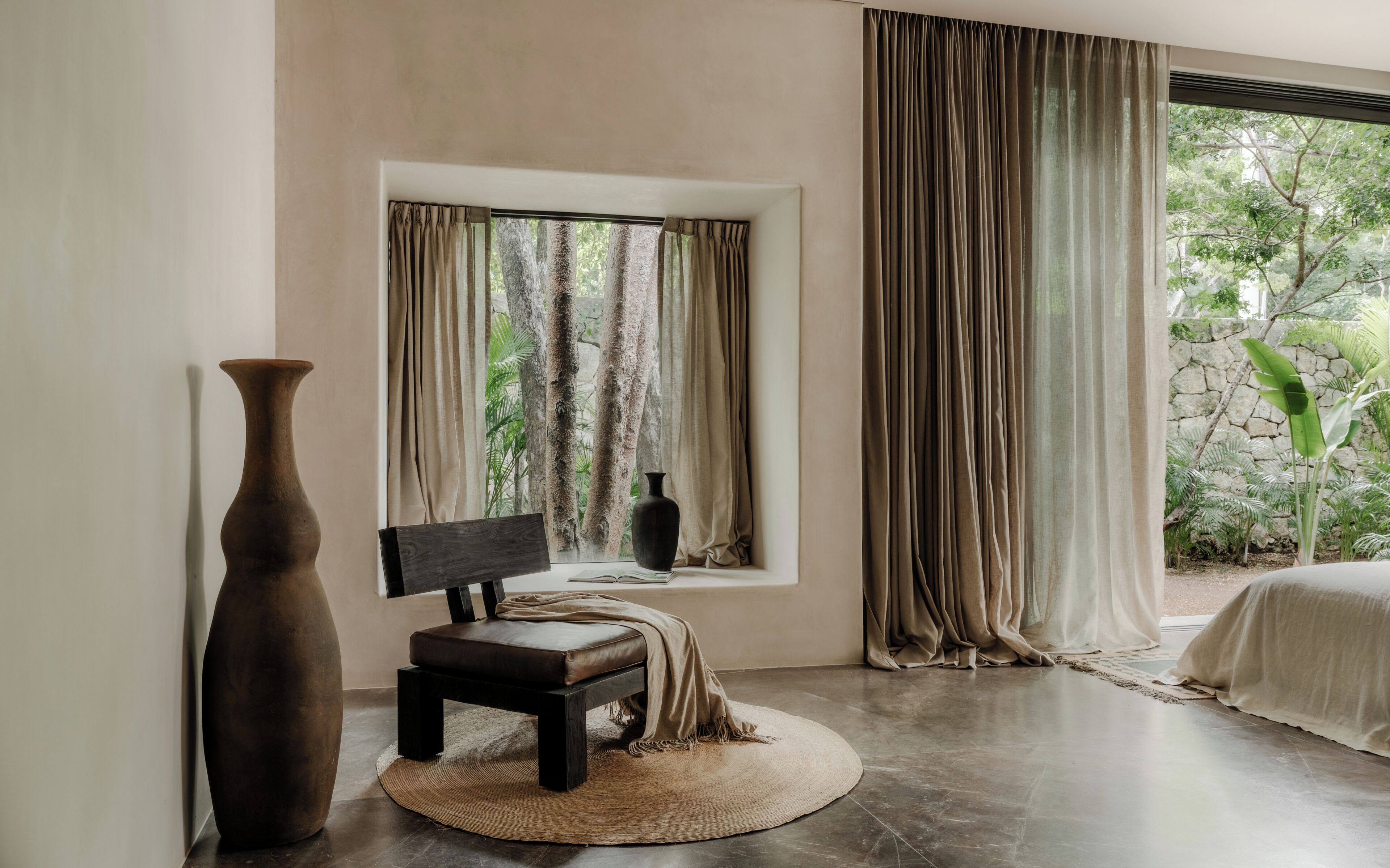 A room with a large window, a bench, a tall vase, and a bed.