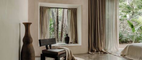 A room with a large window, a bench, a tall vase, and a bed.