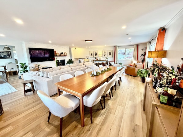 A modern living room with a large wooden dining table, white sofas, and a TV mounted on the wall.