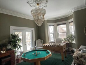 Ein Zimmer mit Billardtisch, Kicker, einer Chandelierleuchtre und zwei Fensterbänken mit Pflanzen.